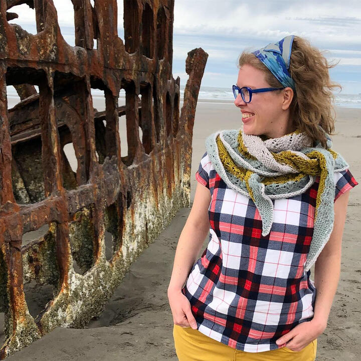 Lee standing on a beach next to an old rusted shipwreck, looking to the side smiling, wearing knit shawl wrapped around neck