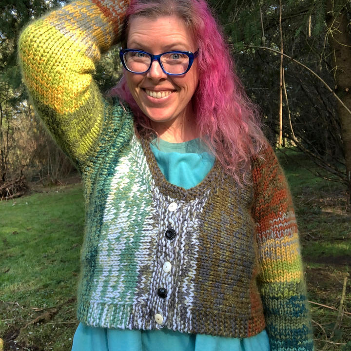 Lee with long magenta hair, hand holding it back, wearing a bulky multi-colored sweater and glasses and smiling