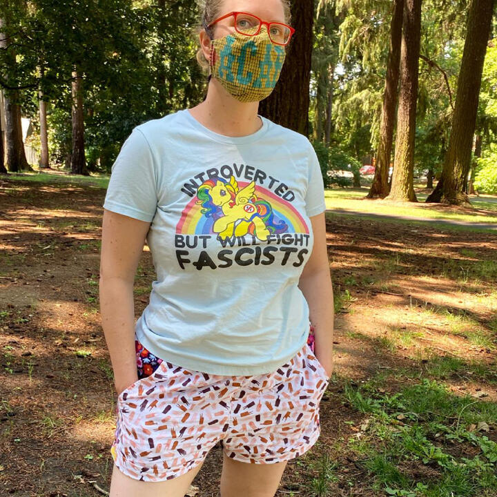 Lee standing in a park, wearing shorts with a print, a light blue tee that says Introverted but will fight fascists, and a mask